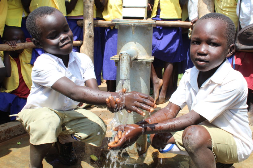 School children in Uganda gather round two boys using a clean water well from Drop in the Bucket