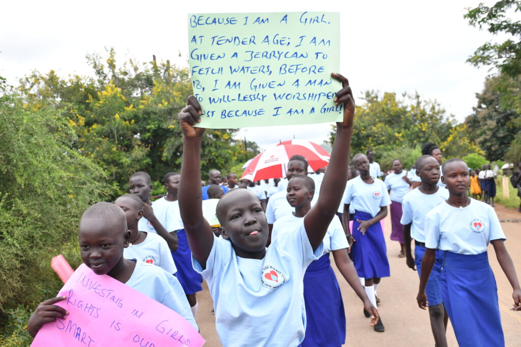 A girl holds a sign "Because I am a girl I am given a jerrycan and told to fetch water"