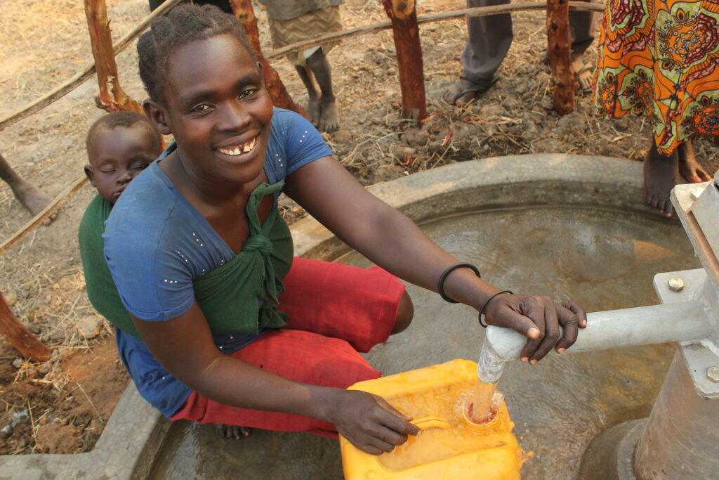 Young mother with a baby on her back collecting clean water from a well drilled by Drop in the Bucket in Opam Okidi North, Amuru, Uganda.