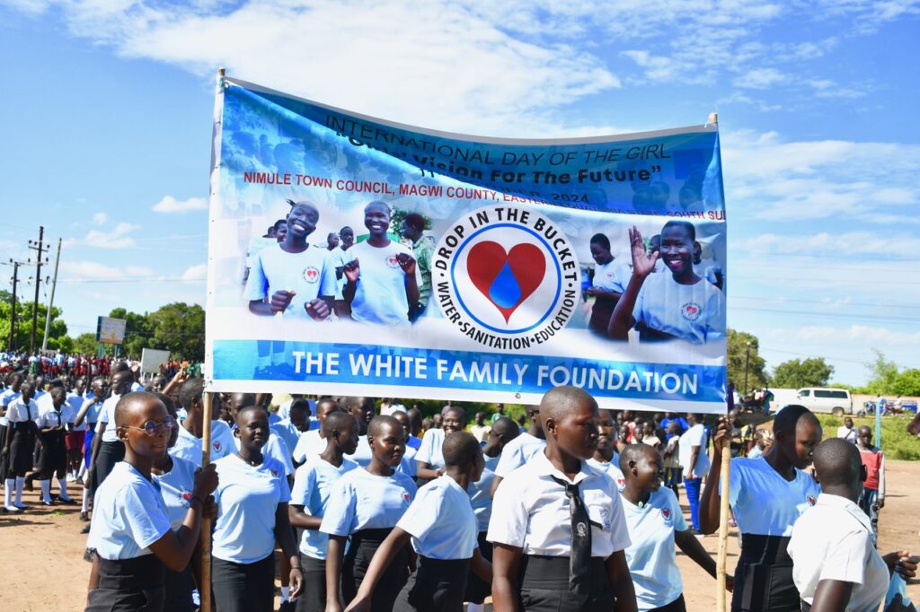 Female students in Nimule, South Sudan, march holding a banner for event sponsor Drop in the Bucket during the International Day of the Girl 2024 celebrations.