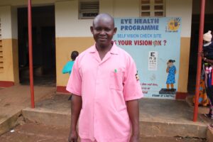 Kamoyi Wilson stands in front of the Naibiri Health Center II where he works as a nurse.