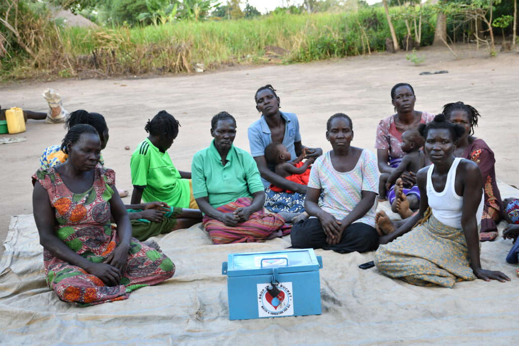 Members of the Bidin B VSLA group gather for a weekly meeting in Nwoya, Uganda. The lockbox in the front of the image bears the logo of the US non-profit Drop in the Bucket